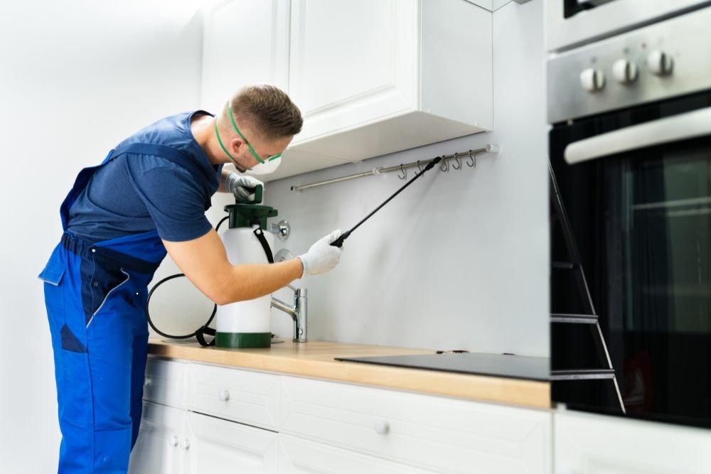 A man in blue overalls uses a spray can in a kitchen, providing ant control services in Renton, WA.